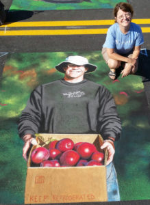 A chalk drawing of a man holding a box of apples and the artist Beth Shistle squatting nearby.